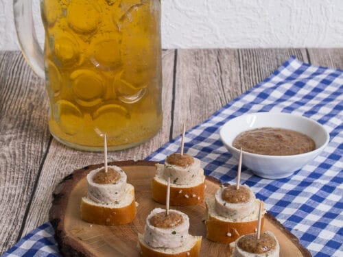 Weißwurst-Häppchen auf Brezelscheiben mit Zahnstochern, serviert mit Senf und einem Bierkrug.