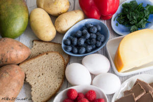 Brot, Kartoffeln, Eier, Beeren, Käse, Paprika, Mango, Grünzeug und Schokolade in einem Arrangement.