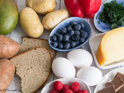 Brot, Kartoffeln, Eier, Beeren, Käse, Paprika, Mango, Grünzeug und Schokolade in einem Arrangement.