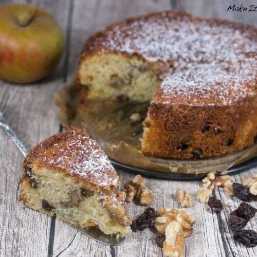 Apfel-Walnuss-Kuchen mit Rosinen 3 Ein Stück Apfel-Walnuss-Kuchen mit Rosinen, mit sichtbaren Apfelstücken und Walnüssen, mit mehr Kuchen im Hintergrund.