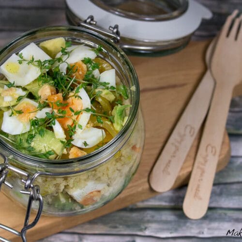 Couscous-Salat im Glas mit Eiern, Kresse und Avocado auf einem Holzbrett mit Besteck.