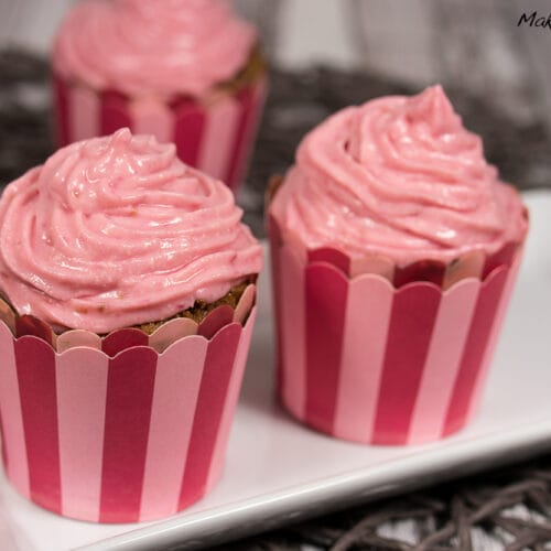 Drei Himbeermuffins mit rosa Topping in gestreiften Verpackungen auf einem weißen Teller.