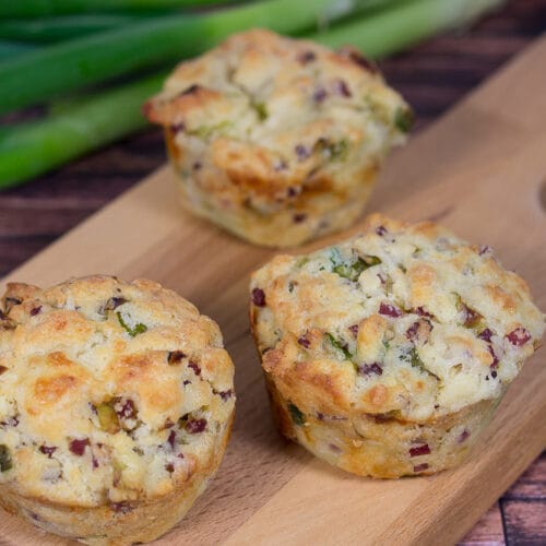 Drei herzhafte Muffins mit Speck und Frühlingszwiebeln auf Holzbrett, weitere Frühlingszwiebeln im Hintergrund.