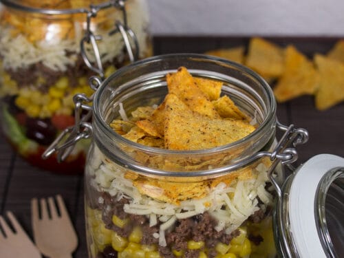 Geschichteter Taco-Salat im Glas mit Tortilla-Chips obenauf, mit sichtbaren Schichten von Salat, Bohnen, Mais und Tomaten.