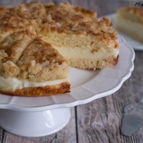 Bienenstich-Kuchen mit Mandeln und cremiger Füllung auf einem Tortenständer, mit einer Scheibe auf einem separaten Teller.