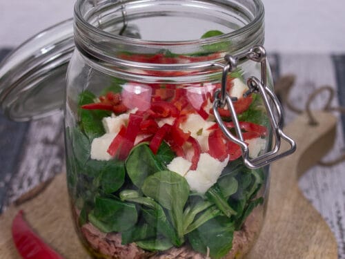 Glas Thunfischsalat mit Mozzarella, Grünzeug und gewürfelter roter Paprika neben einer roten Chili auf einem Holzbrett.