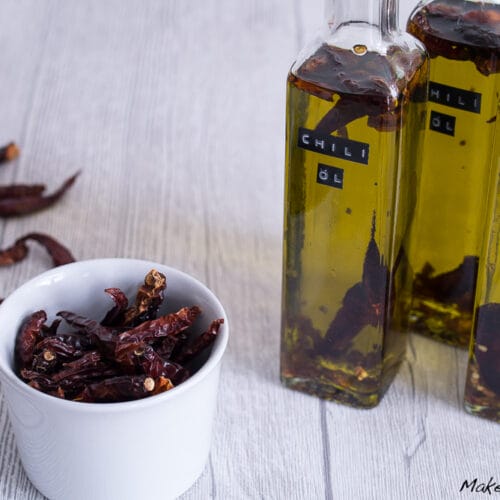 Chiliöl selber machen 3 Drei Flaschen mit Chili-Öl und eine kleine weiße Schale mit getrockneten roten Chilis auf einer hellen Holzfläche.