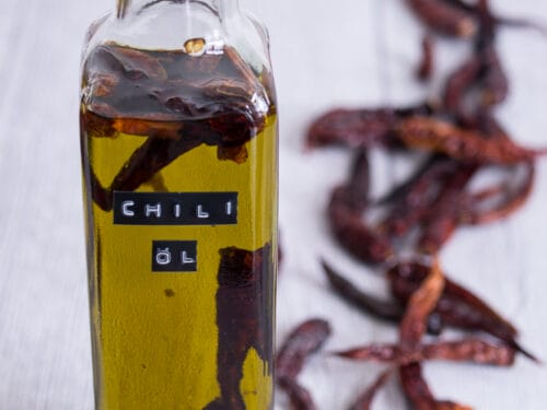 Glasflasche mit Chili-Öl, darin getrocknete Chilischoten, umgeben von ganzen roten Chilis.