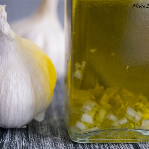 Selbstgemachtes Knoblauchöl 3 Knoblauchzwiebel neben einer Glasflasche mit hausgemachtem Knoblauchöl und gehacktem Knoblauch auf einer grauen Fläche.