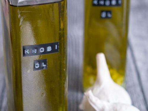 Zwei Glasflaschen mit hausgemachtem Knoblauchöl mit gehacktem Knoblauch und zwei Knoblauchzwiebeln auf einer grauen Fläche.