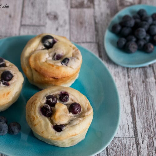 Drei Mini Blaubeerhefeschnecken auf einem blauen Teller, mit frischen Blaubeeren und einer Schale mit zusätzlichen Blaubeeren im Hintergrund.
