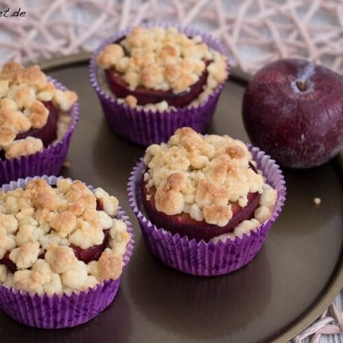 Vier Pflaumen-Muffins mit Streuseln in lila Verpackung und zwei ganze Pflaumen, alles auf einem runden Tablett angerichtet.