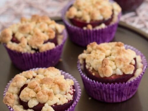 Vier Pflaumenmuffins mit Streusel in violetter Verpackung auf einem Teller, daneben zwei ganze Pflaumen.