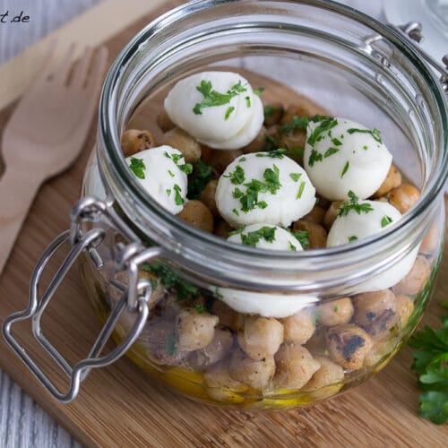 Kichererbsensalat aus dem Glas mit Tomaten, Mozzarellakugeln, Petersilie und Olivenöl auf einem Holzbrett mit einer Holzgabel.