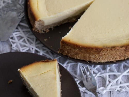 Ein Stück New York Cheesecake liegt auf einem Teller, dahinter der angeschnittene Kuchen.