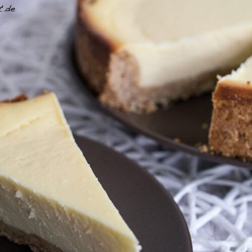 New York Cheesecake 3 Ein Stück New York Cheesecake auf einem Teller, den Rest des Käsekuchens dahinter auf einer Servierplatte.
