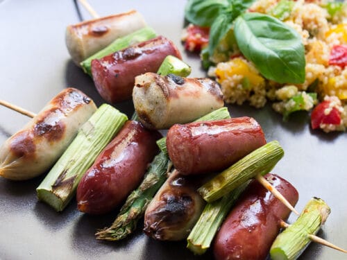 Gegrillte Bratwurstspieße mit Spargel auf einem Teller mit Couscous-Salat und frischem Basilikum.