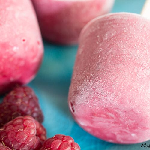 Himbeereis mit Joghurt 3 Rosa Himbeereis mit Joghurt-Popsicles mit frischen Himbeeren auf einer blauen Fläche.