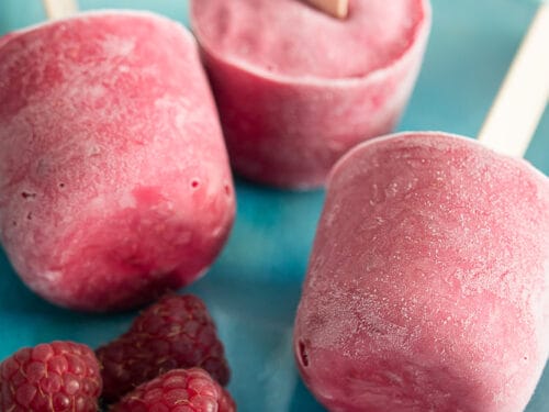 Drei Himbeer-Joghurt-Eis am Stiel mit frischen Himbeeren auf einem blauen Teller.