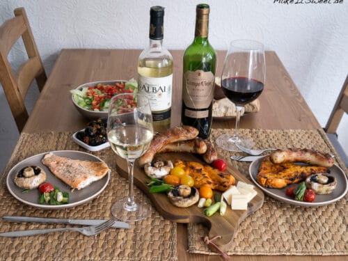 Gegrilltes Fleisch, Gemüse, Salat, Brot, Käse und eine Flasche Rioja-Wein auf einem Holztisch für zwei Personen.