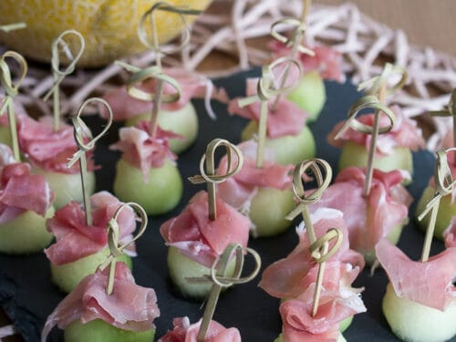 Honigmelone-Kugeln und Prosciutto-Scheiben auf Spießen, angeordnet auf einem Schiefertablett, mit einer halbierten Melone im Hintergrund.
