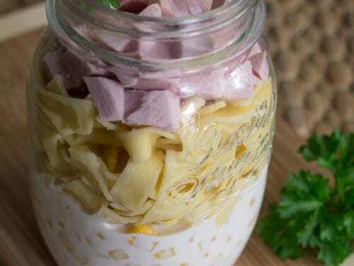 Einmachglas gefüllt mit einer Schicht Joghurtdressing, Mais, Nudeln, Schinkenwürfeln und Petersilie.