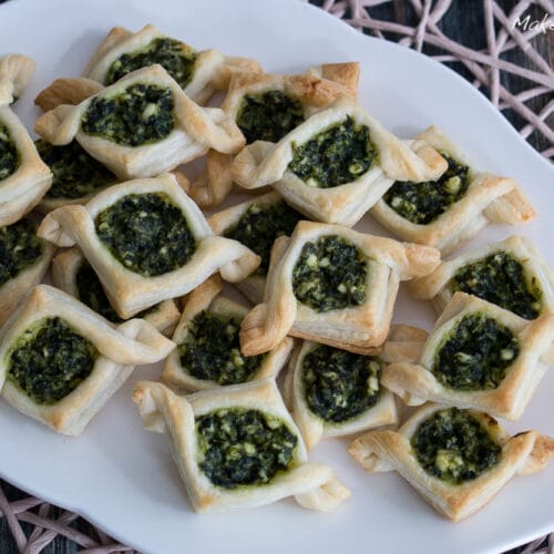 Mini Spinat-Feta-Schiffchen Fingerfood 3 Kleine Blätterteigquadrate, gefüllt mit cremigem Spinat und Feta, auf einer weißen Platte.