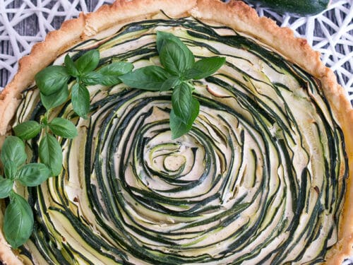 Zucchini-Tarte in Spiralform mit frischem Basilikum und ganzen Zucchini daneben.