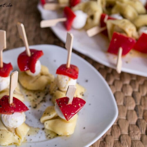 Tortellini Spieße mit Paprika und Mozzarella 3 Tortellini-Spieße mit Mozzarella und Paprika, mit Kräutern bestreut, auf Tellern angerichtet.