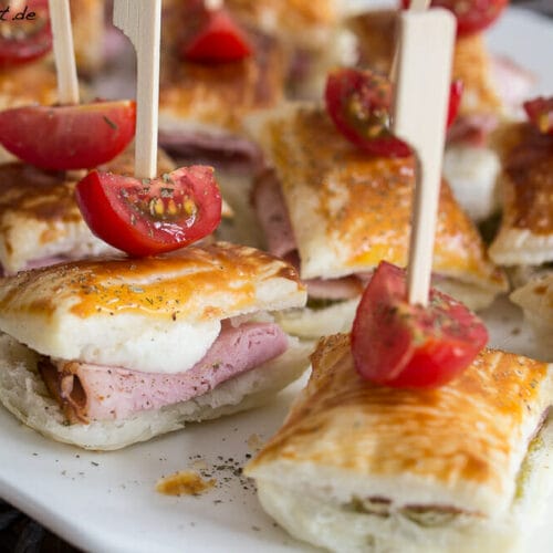 Anzeige - 7 Wonders von Asmodee und Mini Blätterteig Sandwiches 5 Mini-Sandwichspieße aus Blätterteig mit Schinken, Mozzarella, Pesto und Mini-Tomaten auf einem weißen Teller.