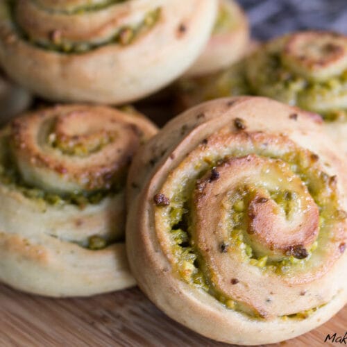 Mini Hefeschnecken mit Pistazien und Zitrone 3 Goldene, spiralförmige Hefeschnecken, gefüllt mit Pistazien, angerichtet auf einem Holzbrett.
