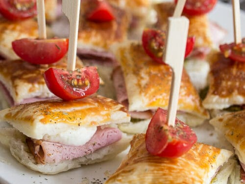 Blätterteig-Häppchen mit Schinken, Kirschtomatenhälften, Pesto, Mozzarella und Holzstäbchen auf einem weißen Teller.