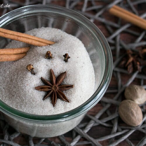 Glasschale gefüllt mit Zucker, Zimtstangen, Nelken und Sternanis für Weihnachtszucker; weitere Gewürze in der Nähe verstreut.