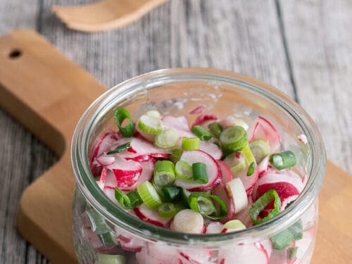 Glasgefäß mit Radieschensalat mit Sahne und Frühlingszwiebeln auf einem Holzbrett.
