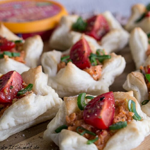 Anzeige - Schnelles Fingerfood mit Gutfried Pulled Chicken Aufstrich 3 Blätterteighäppchen mit Tomate, Frühlingszwiebeln und cremiger Pulled Chicken-Aufstrich-Füllung auf einem Holzbrett.