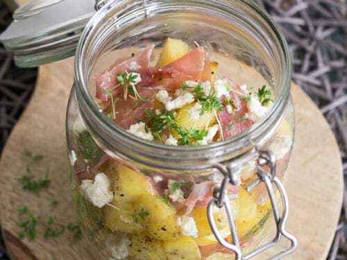 Salat im Glas mit Ofenkartoffel, Prosciutto, Kresse und Feta auf einem Holzbrett.
