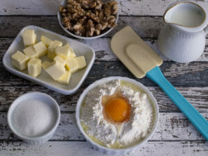 Mehl mit aufgeschlagenem Ei, Zucker, Butterwürfeln, Walnüssen und Milch auf einem rustikalen Tisch mit einem Spatel angerichtet.