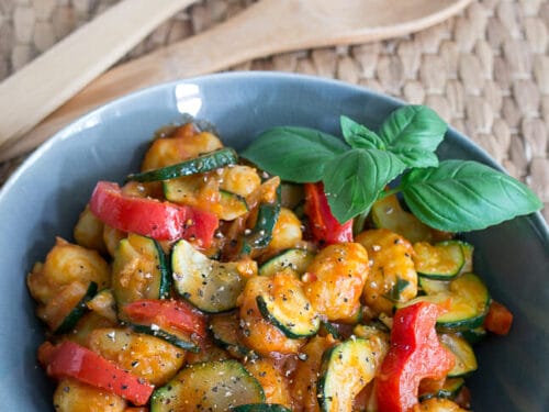 Schüssel mit Gnocchi-Salat mit Paprika, Zucchini, Tomatensauce und frischem Basilikum. Holzutensilien neben der Schüssel.