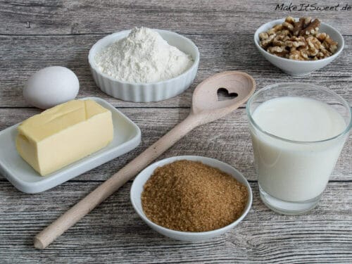 Milch, brauner Zucker, Butter, Ei, Mehl, Walnüsse und ein Holzlöffel liegen auf einem Holztisch zum Backen.