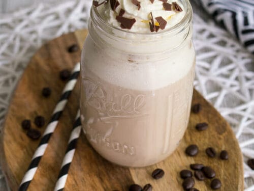 Eiskaffee im Einmachglas mit Schlagsahne und Schokoladenraspeln, umgeben von Kaffeebohnen und Strohhalmen.