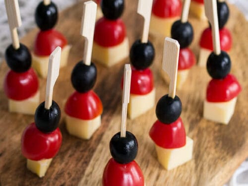 Spieße mit Käse, Kirschtomaten und schwarzen Oliven auf einem Holzbrett angerichtet.