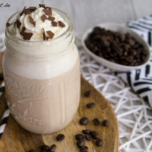 Anzeige- Eiskaffee selber machen und BRITA Tischwasserfilter 7 Eiskaffee in einem Einmachglas mit Schlagsahne und Schokoladenspänen obenauf, neben Kaffeebohnen und Strohhalmen.