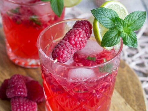 Zwei Gläser Himbeer-Mojito mit Limette, Minze und Eis auf einem Holzbrett, frische Himbeeren an der Seite.