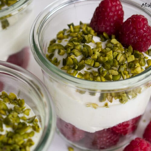 Gläser gefüllt mit Himbeerdessert mit Quark, geschichteten Himbeeren und gehackten Pistazien, gekrönt mit ganzen Himbeeren.