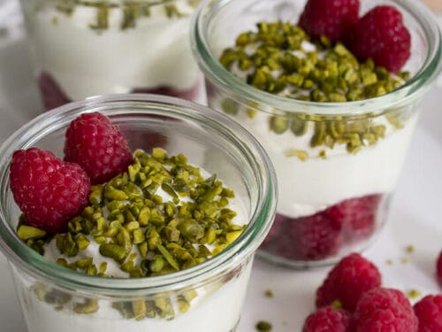 Drei Gläser mit Schichten aus Quark, Himbeeren und gehackten Pistazien, gekrönt mit frischen Himbeeren.