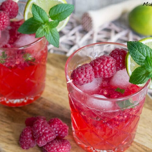 Zwei Gläser Himbeer-Mojito mit Eis, Limettenspalten, Minze und frischen Himbeeren auf einem Holzbrett.