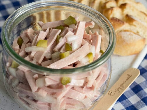 Wurstsalat ohne Käse im Glas mit Essiggurken und Zwiebeln, serviert mit geschnittenem Brot auf einem weißen Teller.