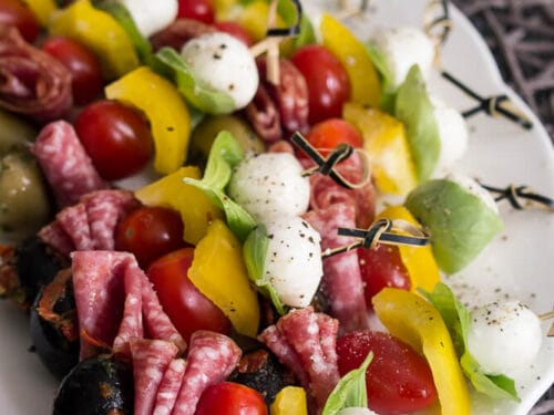 Antipasti Spieße mit Salami, Mozzarella, Tomaten, gelber Paprika, Oliven und Basilikum auf einem weißen Teller.