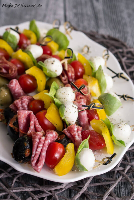 Antipasti Spieße auf einem Teller mit Oliven, Salami, Tomate, Paprika, Mozzarella und Basilikum