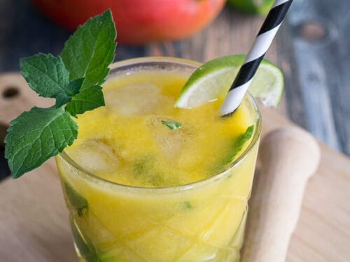 Gelber Mango-Mojito mit Eis, Minze und einer Limettenspalte auf einem Holzbrett; frische Mango und Limette ebenfalls abgebildet.